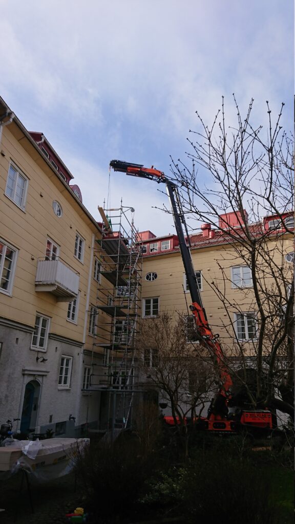 Jekkokran på en innergård mellan gula hus, med kranarmen upp mot taket intill träd och byggställning.